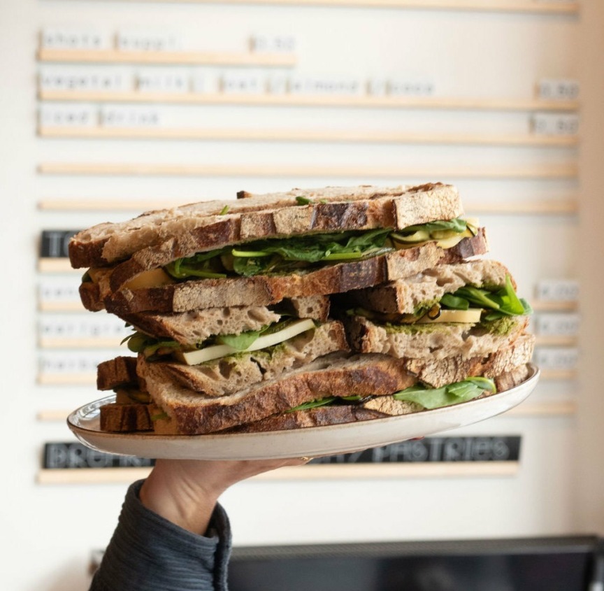 A person holds a plate with a tall stack of sandwiches filled with fresh greens and cheese. The background shows a blurred menu.