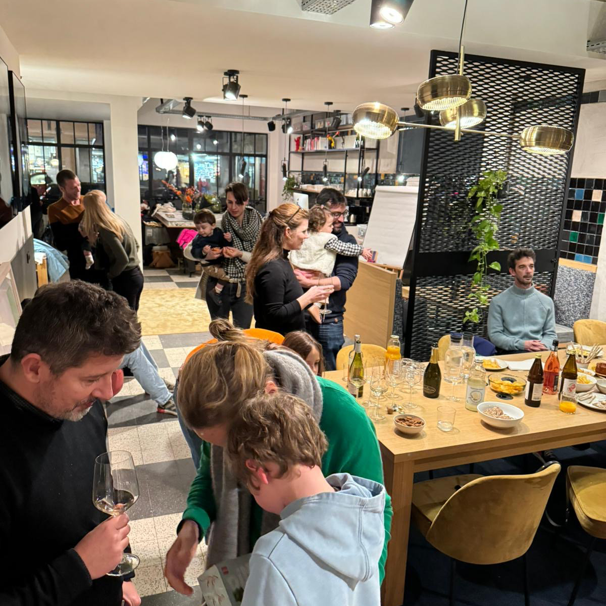 A group of adults and children are gathered in a cozy, modern kitchen. Some are chatting and holding drinks, while one person reads with a child. The atmosphere is warm and lively.