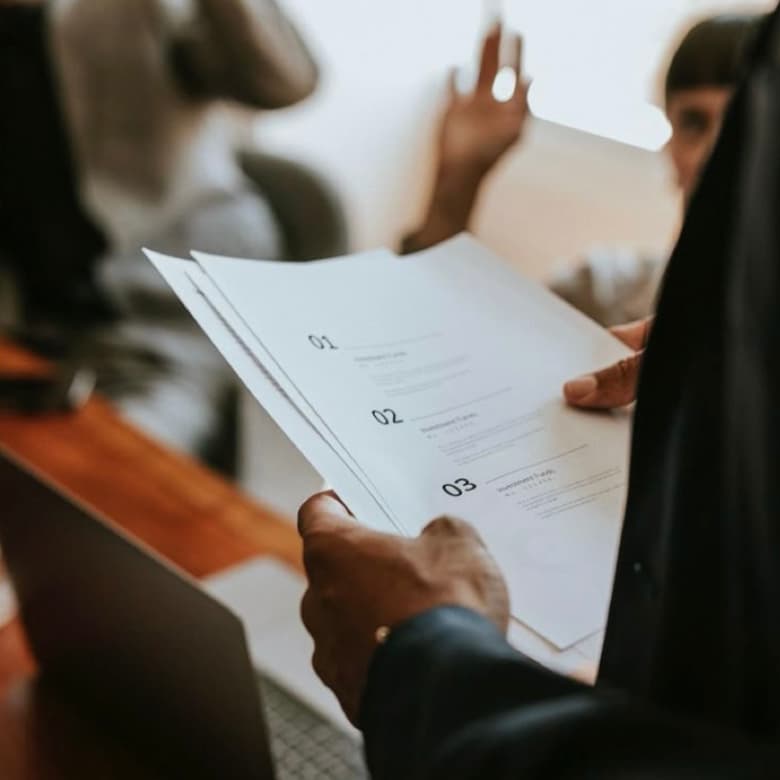 A person in a business suit holds documents numbered 01 to 03. In the background, another person raises a hand, suggesting a meeting or discussion.