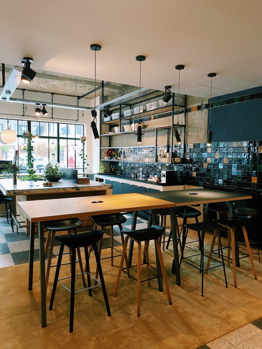 Modern kitchen with hanging lights, wooden tables, and high stools. Black tiled backsplash, shelves with plants, and large windows, creating a warm atmosphere.