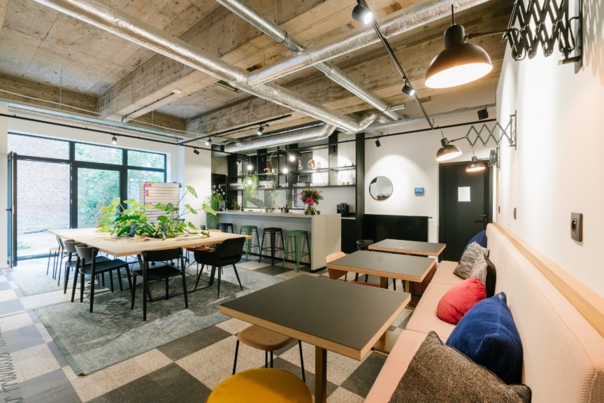 Modern coworking space with communal table, plants, sofa seating, and industrial-style lighting.