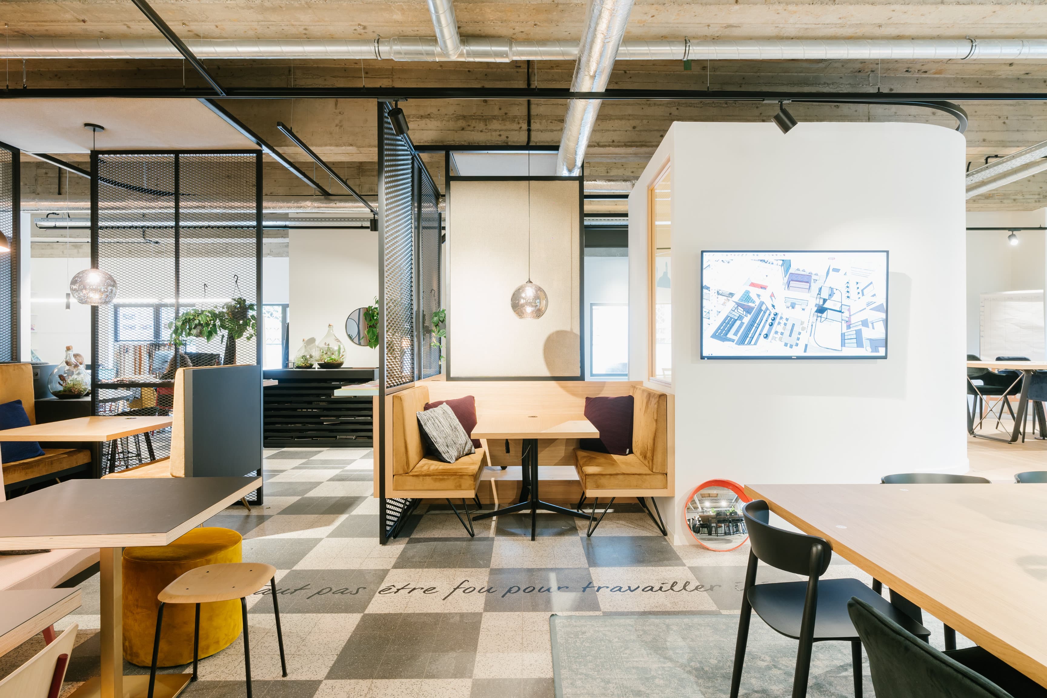 Modern coworking space with cozy seating, pendant lighting, and a wall-mounted screen. Open design with light wood and neutral tones.