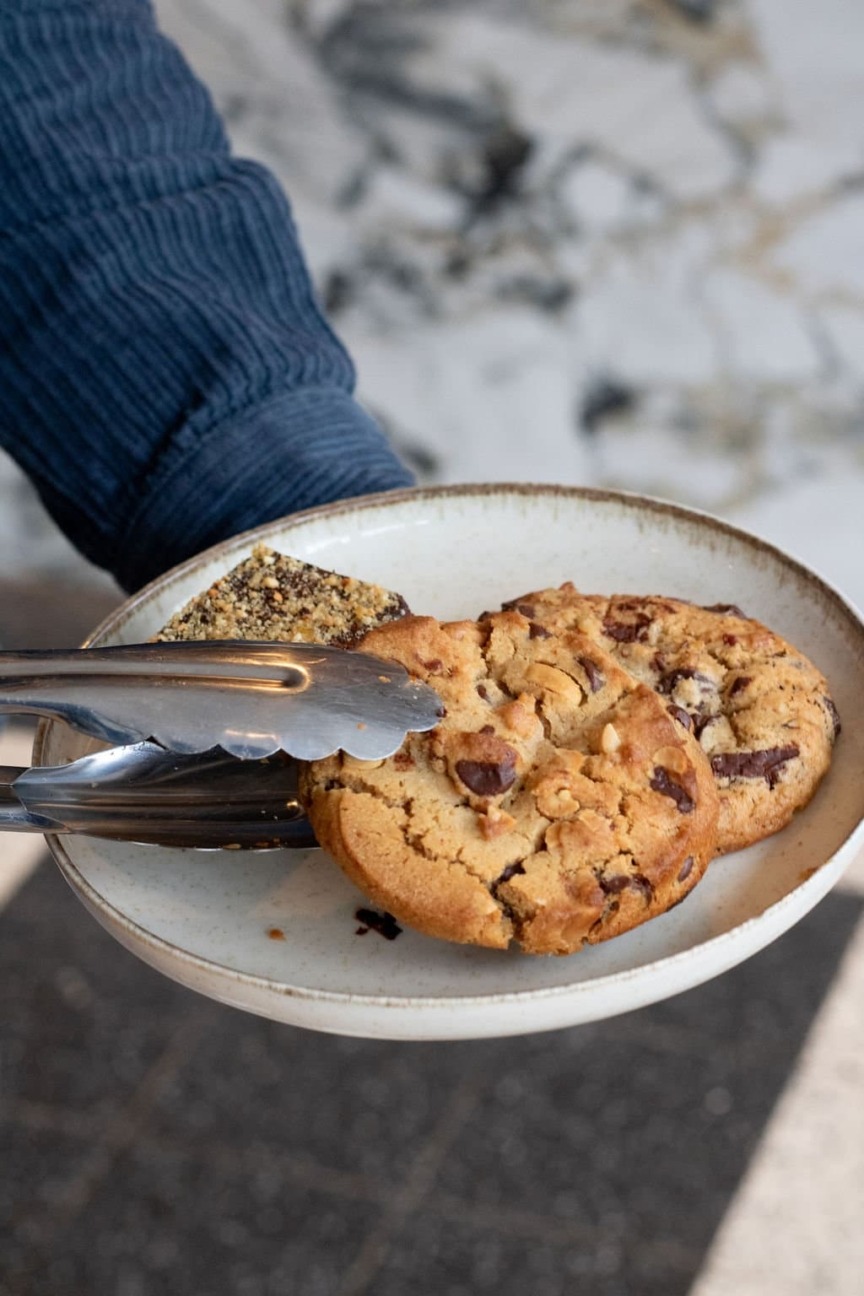 A person in a blue sleeve holds a plate with chocolate chip cookies and a textured bar, using metal tongs. The setting is cozy and inviting.