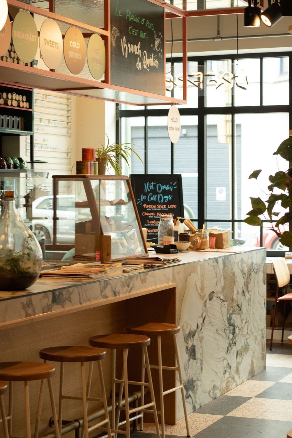 Cozy café interior with a marble countertop, wooden stools, and a blackboard menu. Warm lighting creates an inviting atmosphere. Plants and decor add charm.