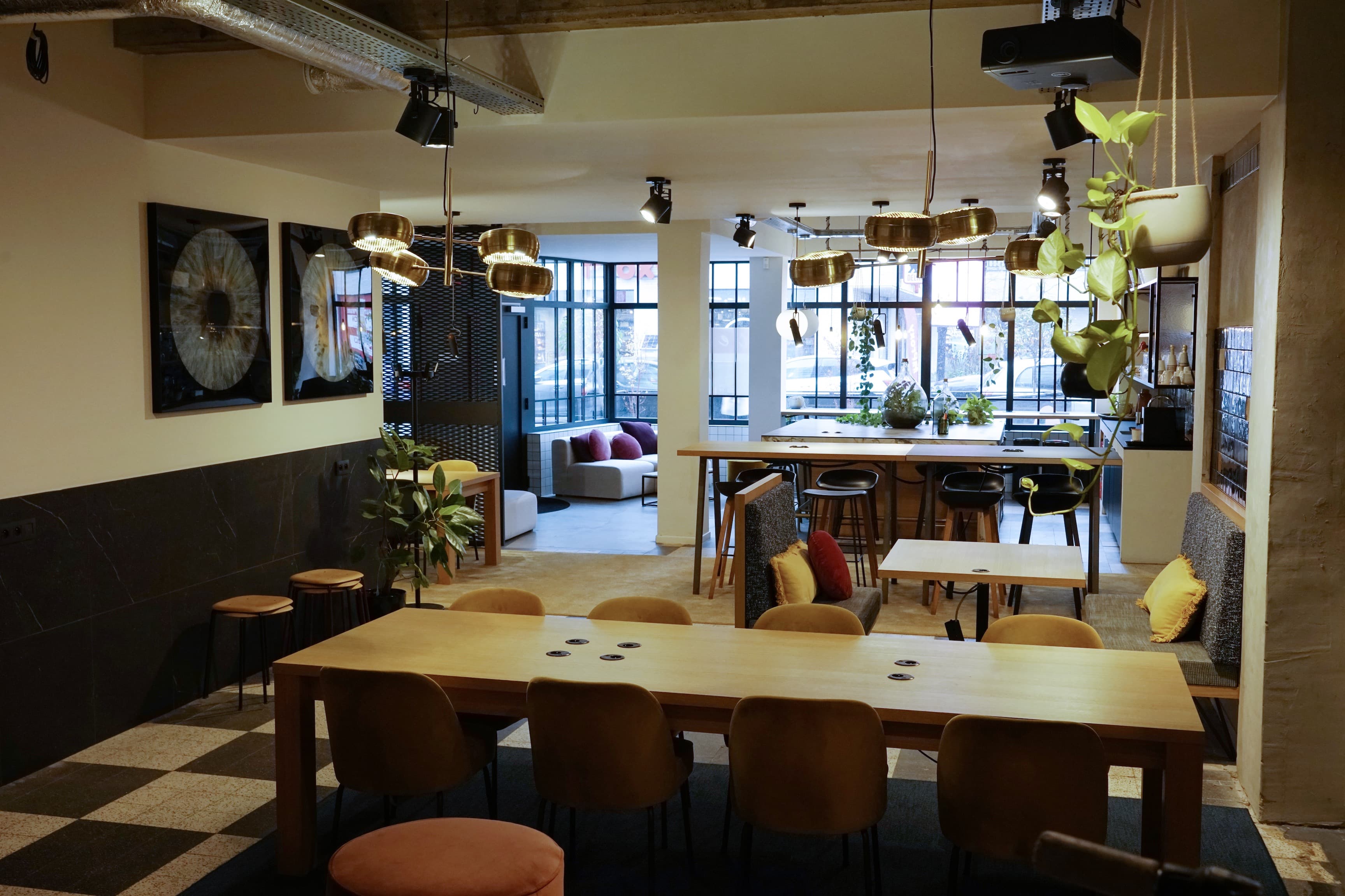 A cozy co-working space with a wooden table and cushioned chairs. Hanging plants and pendant lights create a warm atmosphere. Large windows allow natural light in.