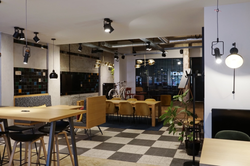 A modern, stylish cafe interior featuring wooden tables, contemporary pendant lights, and checkered flooring. A bicycle decorates the back, adding charm.