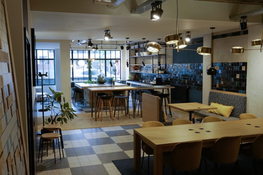 Stylish, contemporary café interior with wooden tables, cushioned chairs, and a checkered tile floor. Pendant lights hang above, adding a warm glow.